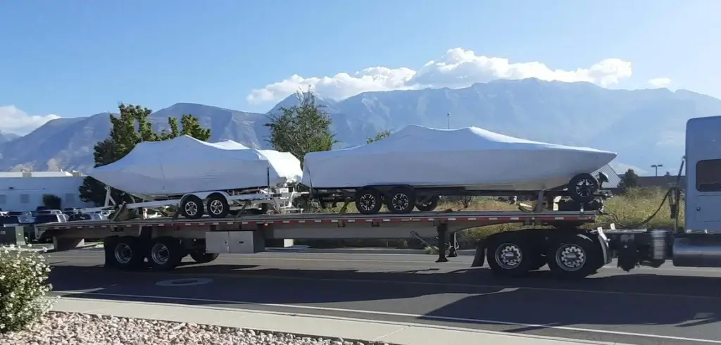 Flatbed truck transporting two covered boats on trailers with mountains in the background