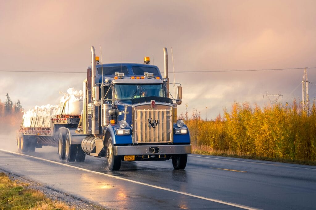 A blue semi-truck hauling a covered load down a wet highway